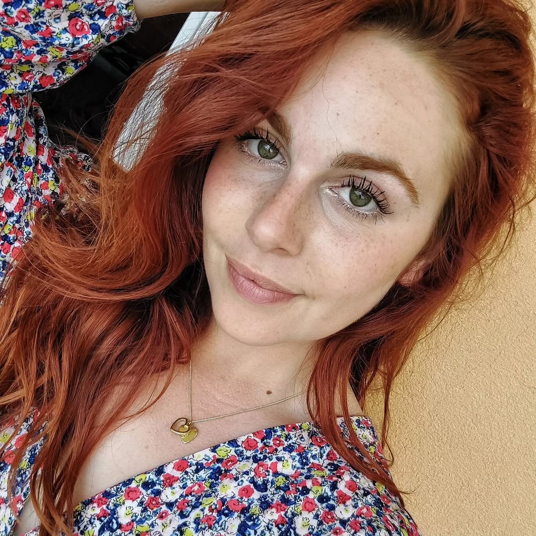 Chic Summer Red Hair Looks 2026: Casual Style A woman with radiant copper red hair smiles, wearing a white oversized linen shirt, blue denim shorts, and sunglasses, embodying cool summer red hair looks 2026.
