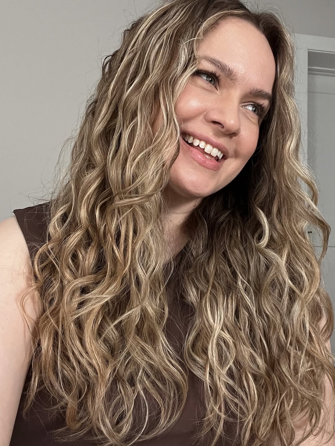 Beautiful woman with naturally curly brown hair styled in soft, bouncy waves, perfect for summer hair curly ideas 2026. She wears a relaxed off-white linen top and looks effortlessly chic for a casual day out.