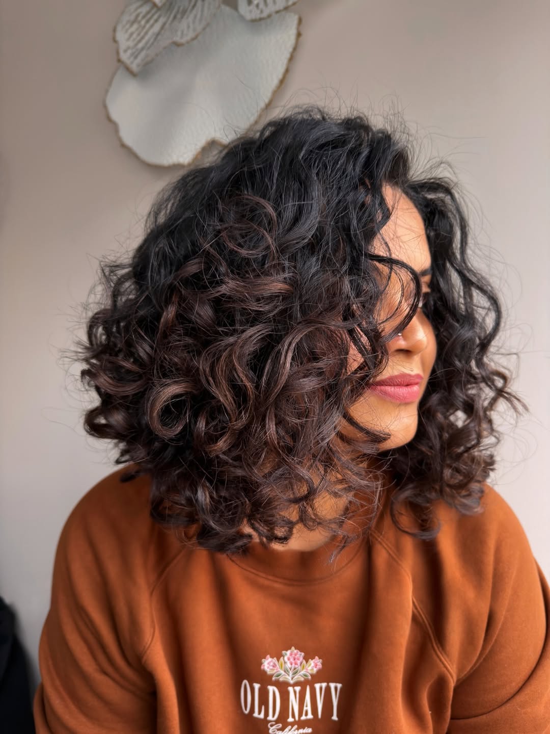 Beautiful woman with defined, voluminous summer hair curly ideas 2026, styled in natural waves, sitting outdoors casually. Her dark curls cascade over her shoulders, highlighting a relaxed summer vibe.