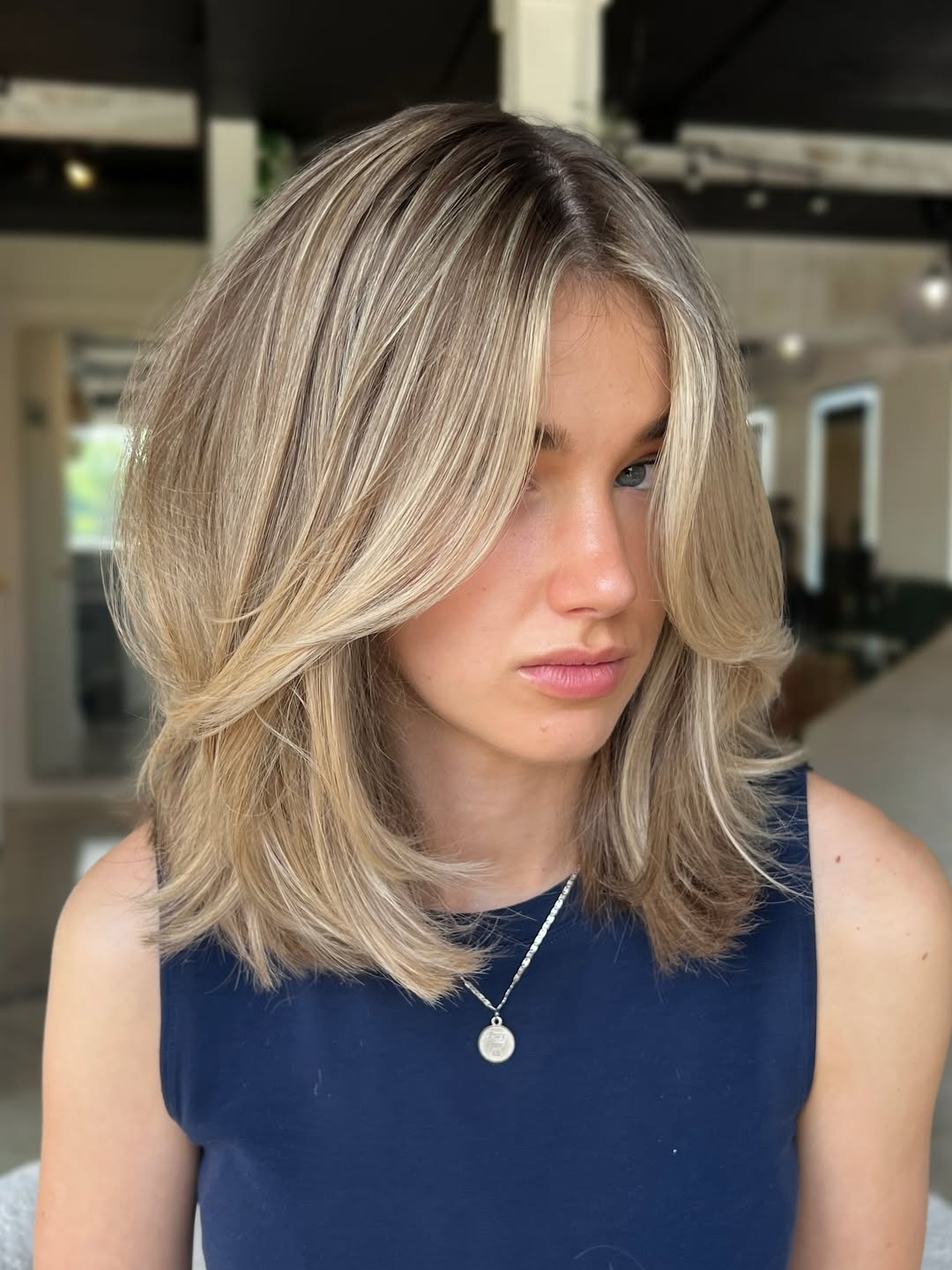 A woman confidently poses outdoors, her long, wavy brown hair showcasing a popular summer hair trend 2026. She wears a chic white off-the-shoulder top and blue high-waisted denim jeans, perfect for casual summer outings.